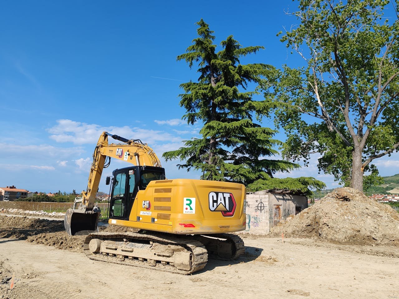 Home A CAT excavator working at a construction site with trees and blue sky in the background.